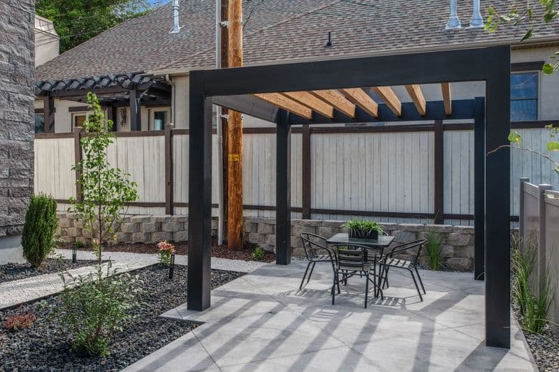 Modern patio with a pergola, table, and chairs surrounded by stone landscaping and greenery.