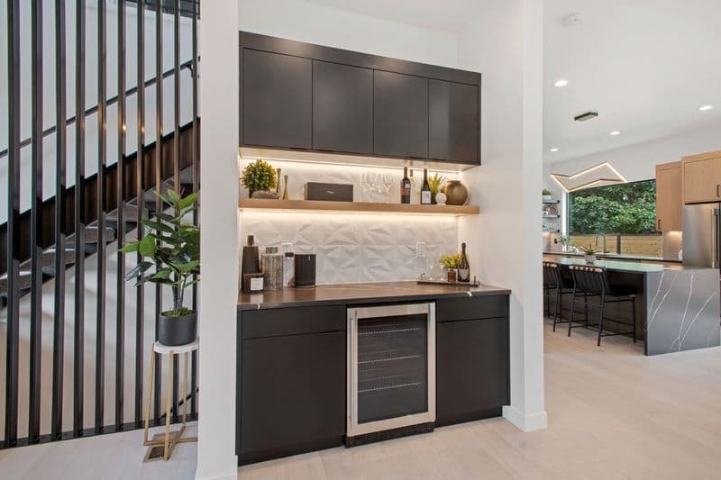 Modern kitchen wet bar with black cabinetry, shelves, and wine fridge in bright open space.