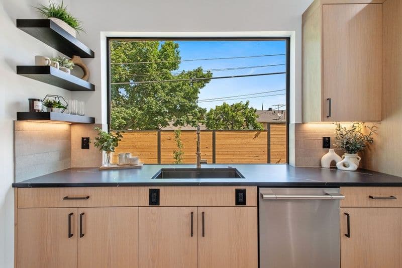 Modern kitchen with wooden cabinets, sleek sink, and large window showcasing green outdoors.